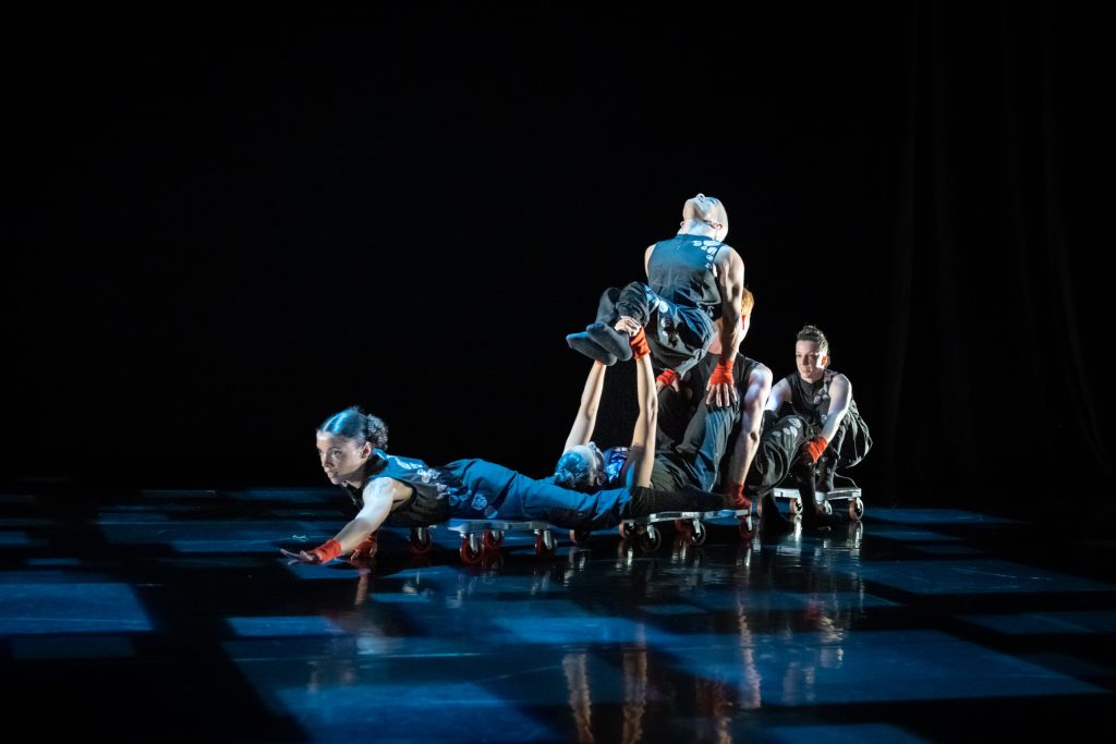 A photo of the AXIS dancers traveling across the floor on wheeled scooterboards in an acrobatic cluster of bodies. 