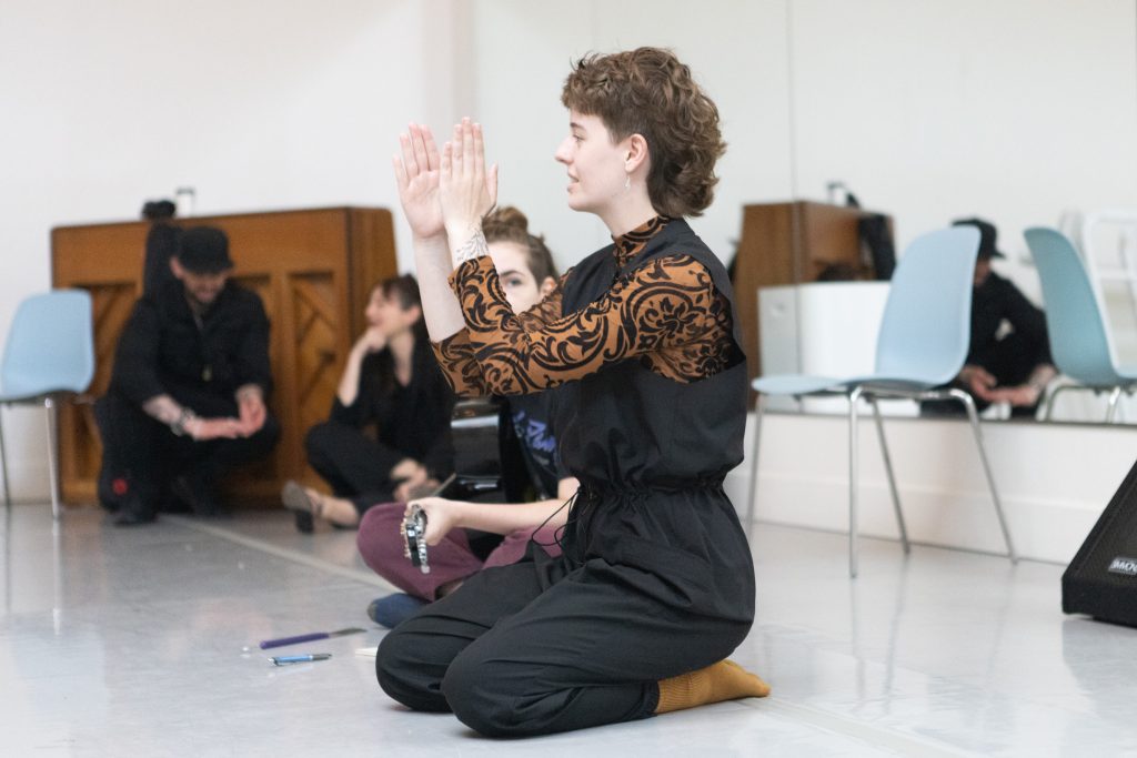 Sammie raises two upper limbs while directing dancers in rehearsal. Have brown wavey hair cut into a mullet, wearing a black jumpsuit and brown socks and turtleneck.