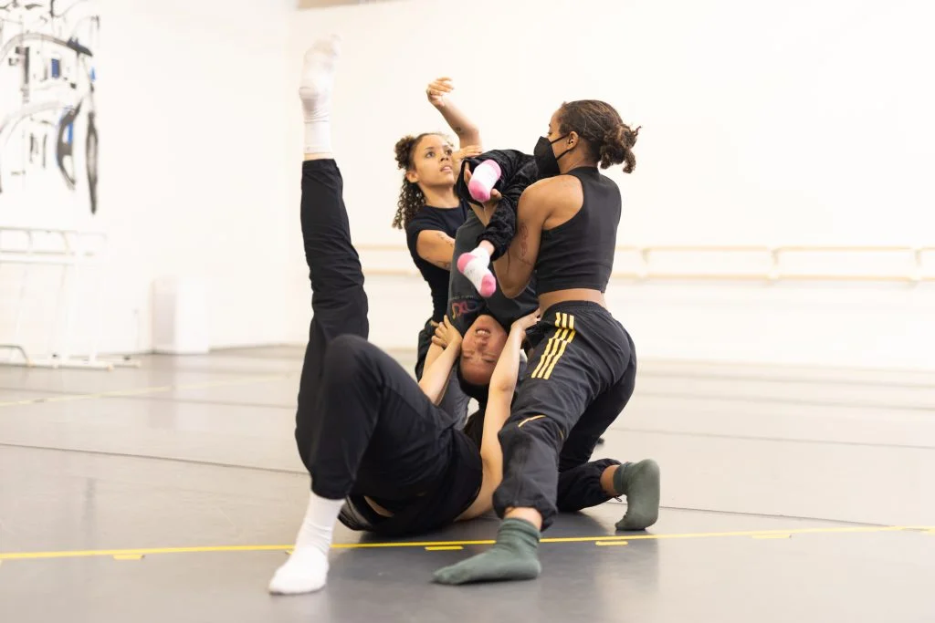 In work by Saira Barbaric, Alaja and Zaria kneel and flip Julie upside down while Zara lifts Julie from Julie's shoulders.