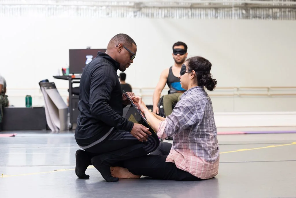 DJ and Zara talk and use touch to communicate in the dance studio with a black binder. DJ, who is blind, wears a black track suit and sunglasses. Zara and Janpi wear black eye masks that cover their faces.