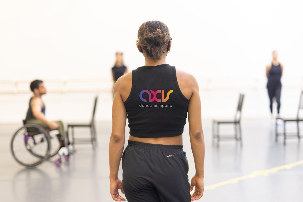 Zaira from the back in rehearsal. They wear a black top with the rainbow gradient AXIS logo.