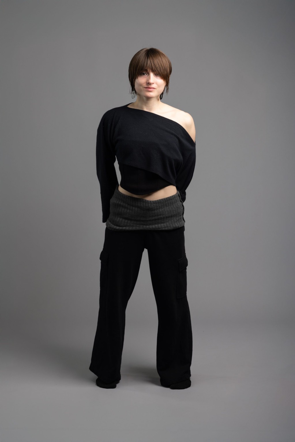 A portrait of Anna, who looks confidently at the camera. She has short-brown hair in an angular pixie-bob, long textured oblong earrings, and an asymmetrical black black shirt revealing the skin of her shoulders on her left side.
