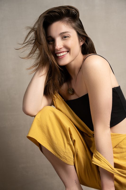 Anna, a young white woman in a yellow jumpsuit, sits casually on a stool with her long brown hair tousled over her right shoulder. She smiles warmly at the camera.