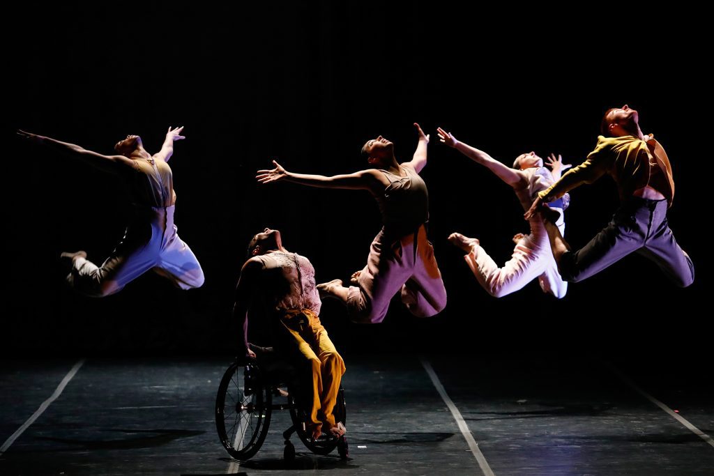 Five disabled and non-disabled dancers jump in a line. Four dancers leap in the air with arms outstretched, one dancer pushes themselves up out of their wheelchair.