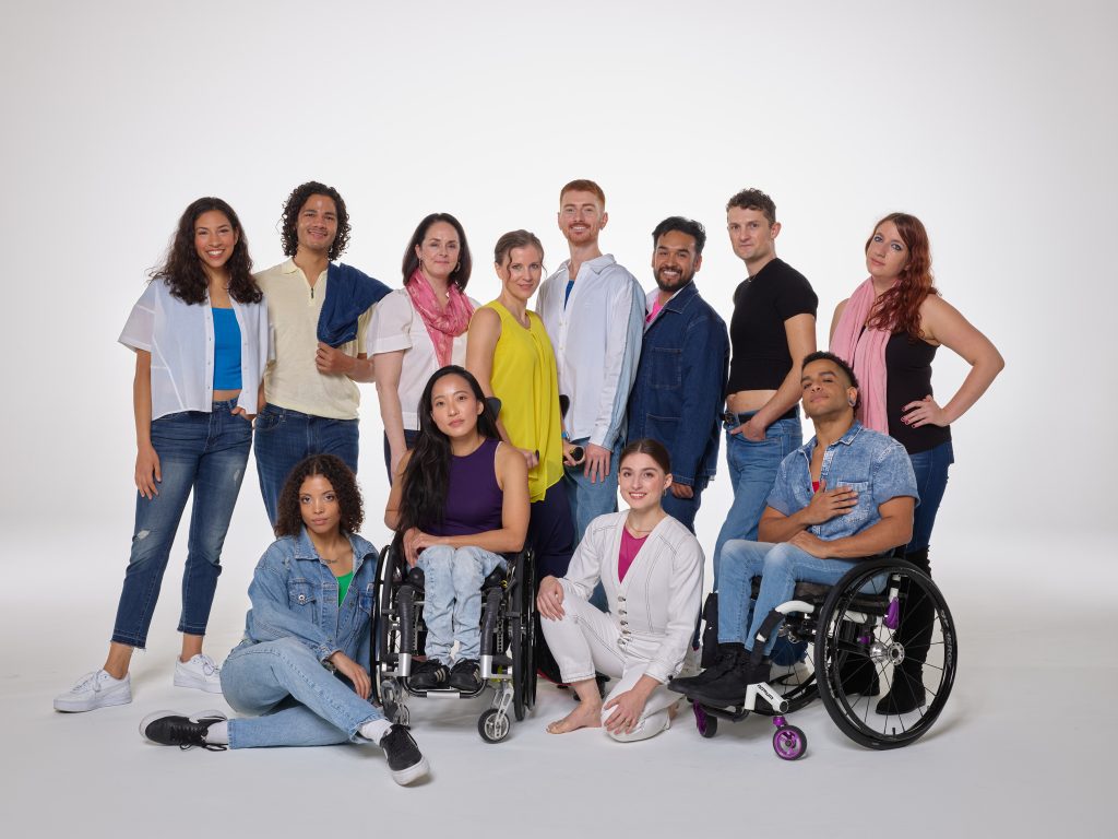 A group of twelve disabled and non-disabled AXIS dancers and staff pose together against a white background, wearing denim with blue, pink, yellow and white accents. 