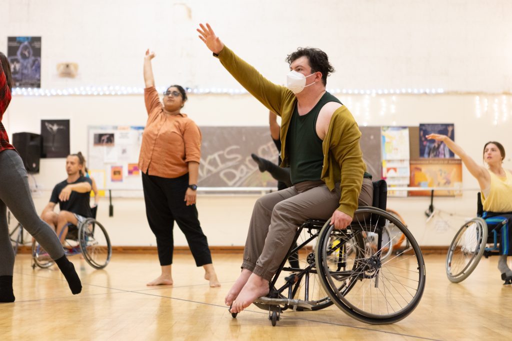 A diverse group of disabled and non-disabled dancers take class together; lifting one upper limb forward in space as they watch an instructor who is out of the frame. Some dancers are using wheelchairs and others are standing.