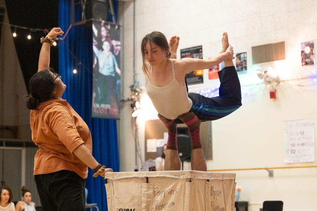 Three dancers perform a group exercise during the intensive. One dancer lies in a big laundry bin, lifting another in the air 'airplane style' while a third dancer draws a line upwards.