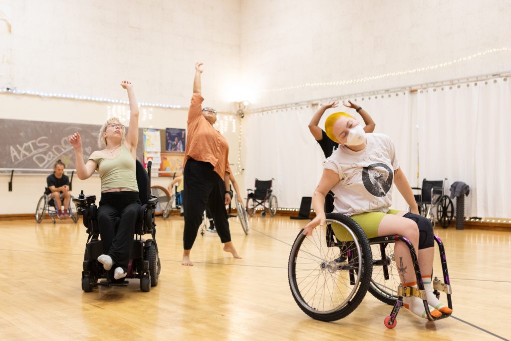 A diverse group of disabled and non-disabled dancers take class together; reaching to the ceiling with one or two upper limbs. Some dancers are using wheelchairs and others are standing.