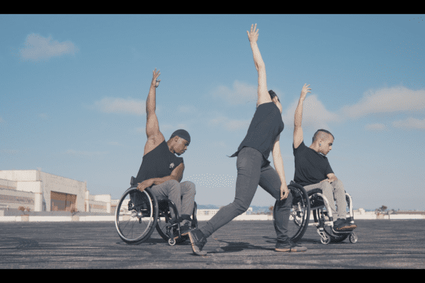 Three disabled and non-disabled dancers reach one limb up to the sky, turning their faces to profile. They wear all black, and perform outside in a large parking lot by the San Francisco bay.