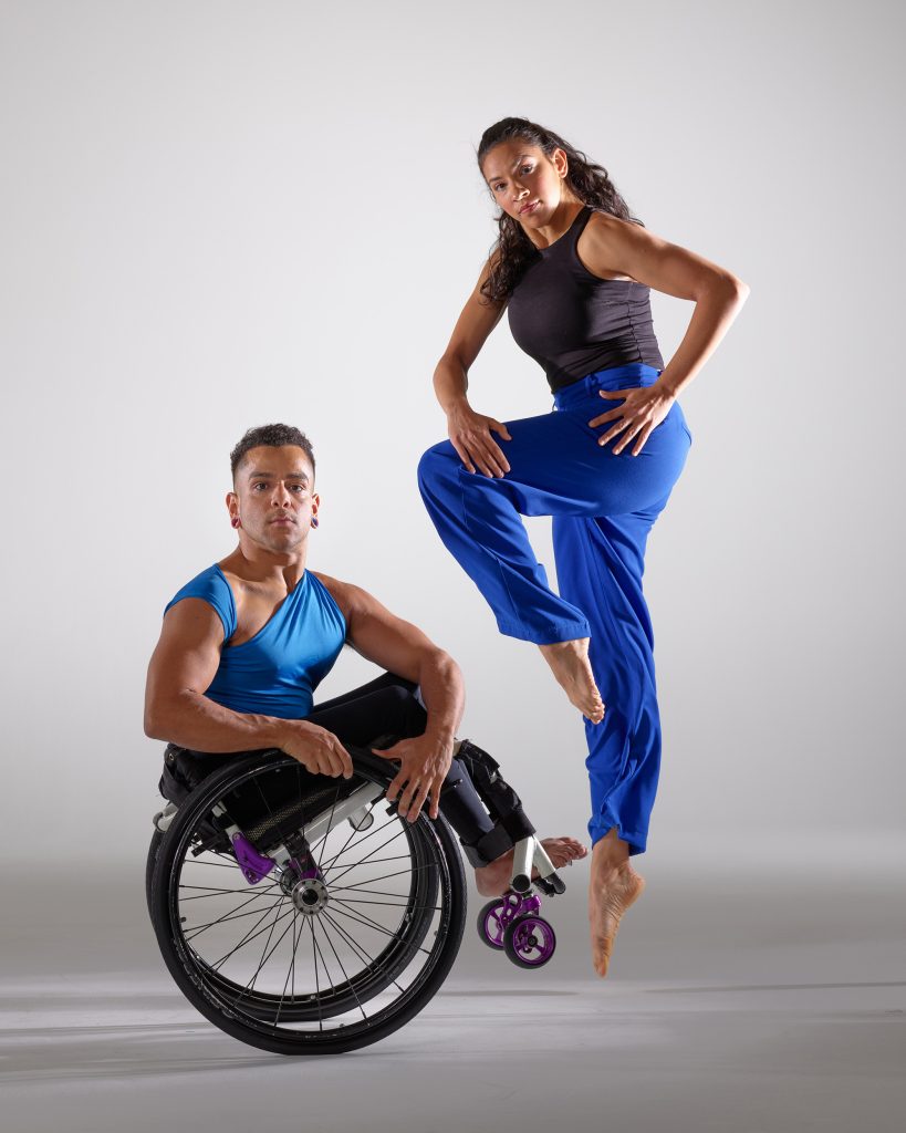 Janpi holds a wheelie in their wheel chair, cheating their torso to the camera. Zara holds a similar position with her hands as she jumps upwards with one leg bent at the knee. Both dancers wear blue and black costumes.
