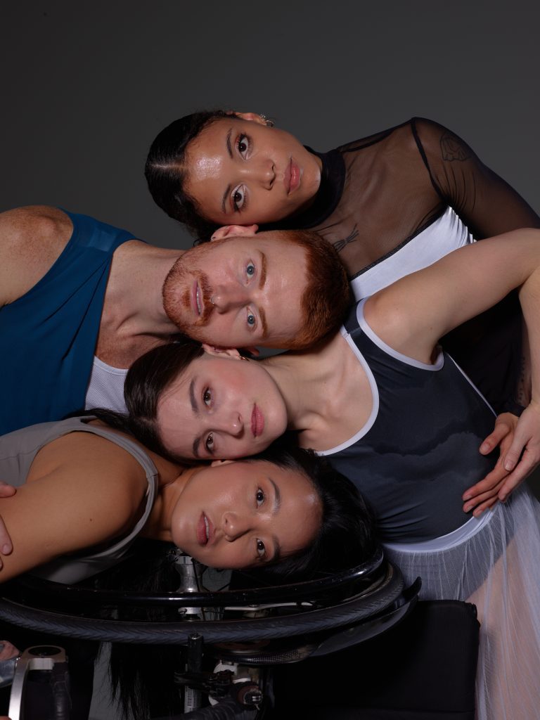 Four diverse dancers rest their heads again each other with intimacy in a vertical column. The dancers on the bottom, Julie Hasushi, rests her head on her wheelchair.