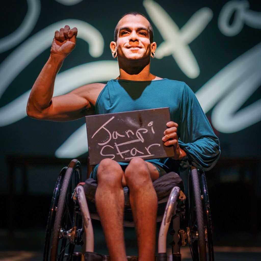 JanpiStar, who is a wheelchair user, signs their name on stage while holding a chalkboard with their name written on it.