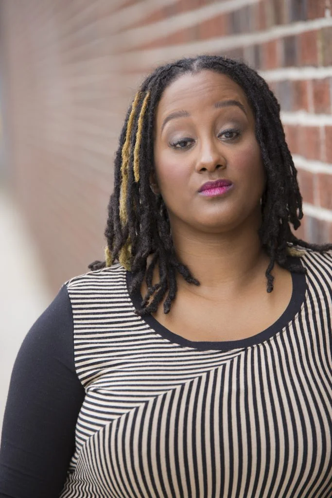 This is a headshot of Kayla Hamilton, who is a dark brown-skinned Black woman. She is posing in front of a blurred brick wall. She is wearing a long sleeve black & tan striped shirt. She has light makeup and her gaze is towards us. Her black & golden highlighted dreads are down.