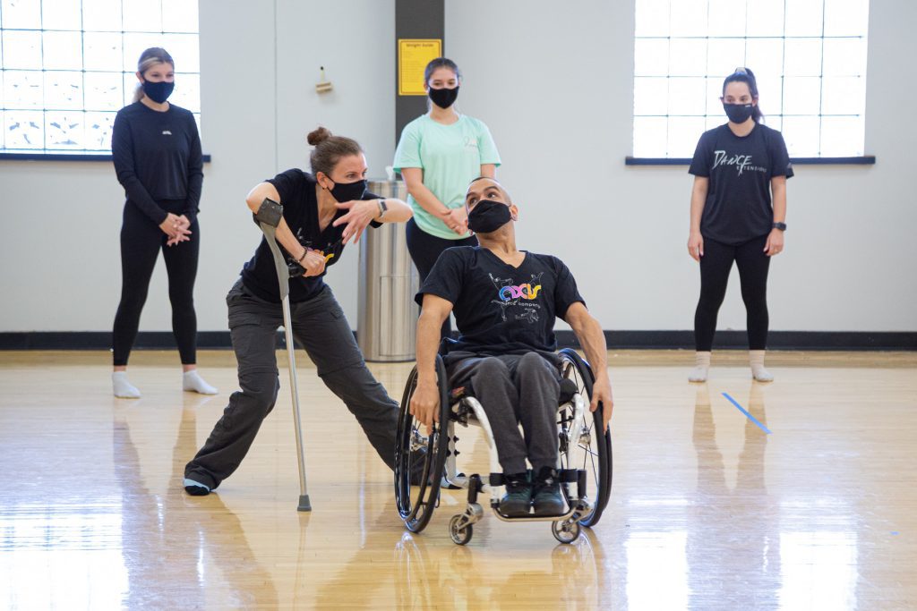 Nadia Adame and JanpiStar demonstrate an improvisation together while a group of dancers watch. Janpi leans back in their wheelchair while Nadia reaches out with one arm and leg, balancing on her cane with her knee turned inwards.