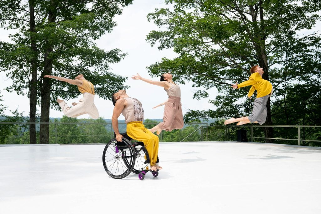 A diverse group of disabled and non-disabled dancers perform on a white outdoor stage, which opens up to a majestic background of trees, rolling hills and sky. JanpiStar lifts their torso out of their wheelchair in a high backward arch. Alaja, Zara, Louisa and David translate this gesture as they leap into the air; chests out with lower and upper limbs reaching backward. The dancers wear flowing costumes in shades of yellow, grey, and beige