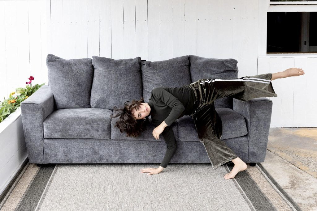 Clairey, a short, white woman is pictured catching herself falling horizontally off of a grey couch. One of her hands and one of her feet make contact with the ground. Her other foot is outstretched, her other hand in motion near her face. She is wearing a dark green shirt and velvety dark green pants. Her hair is tousled. In the background, there is a white fence, a flower bed, and a window into a dark room.