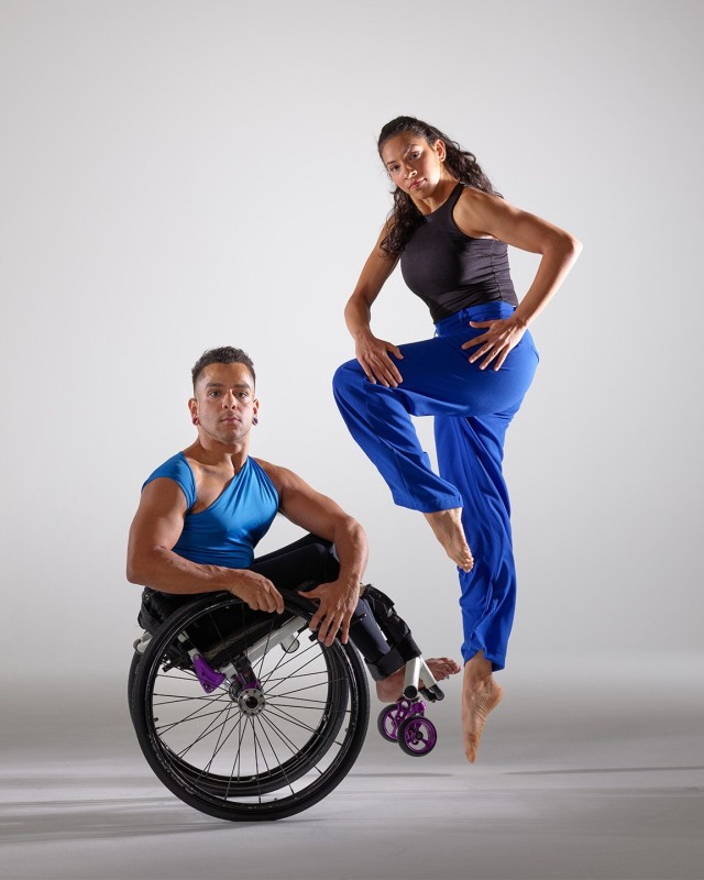 Janpistar holds a wheelie in their wheelchair, twisting their torso forward, while Zara jumps in the air with one foot held up and holding two arms forward with bent elbows. The dancers wear blue costumes on a white background.