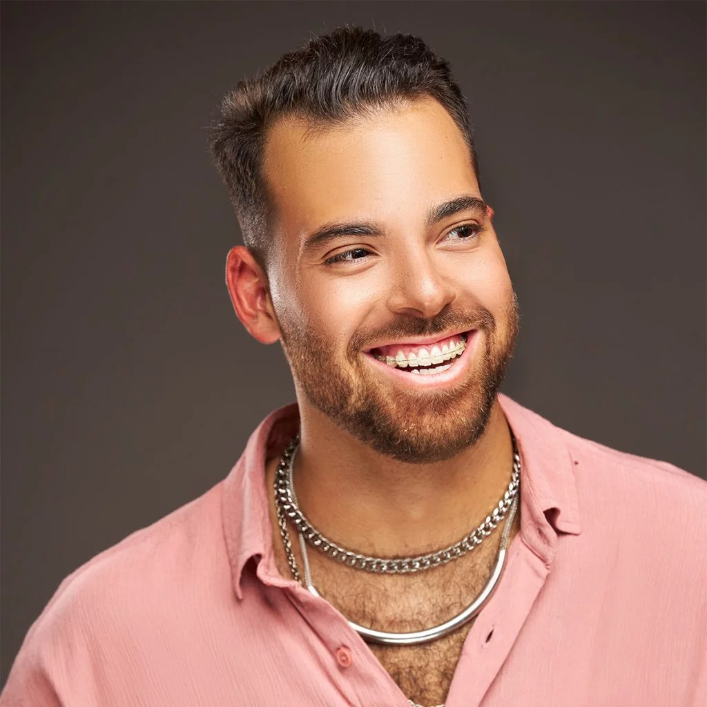 Brian smiles for the camera; he has light tan skin, brown hair and beard; wearing a pink shirt and silver chains.