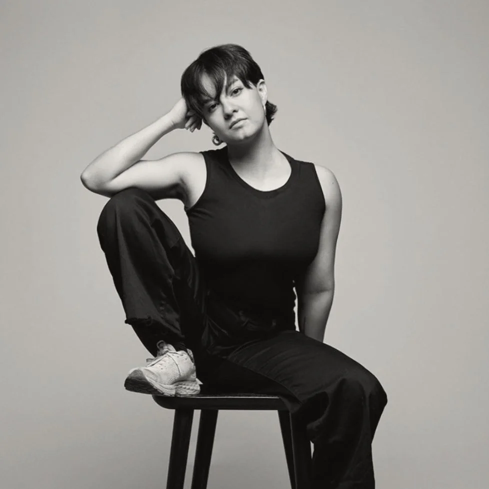 Joelle poses with a focused gaze, hand supporting her head, wearing a black tank top.