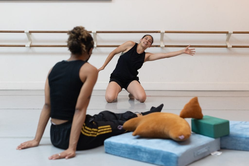 Dancer z sits on the ground next to an assortment of floor pillows, while Zara kneels and stretches her hands on a horizontal plane.