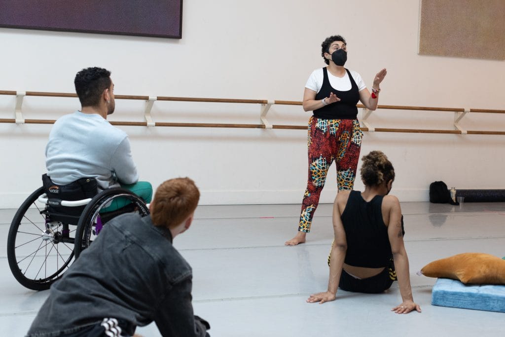 Choreographer Larissa Velez-Jackson stands, wearing red patterned pants and a black face mask, speaking to three disabled and non-disabled artists in the studio.