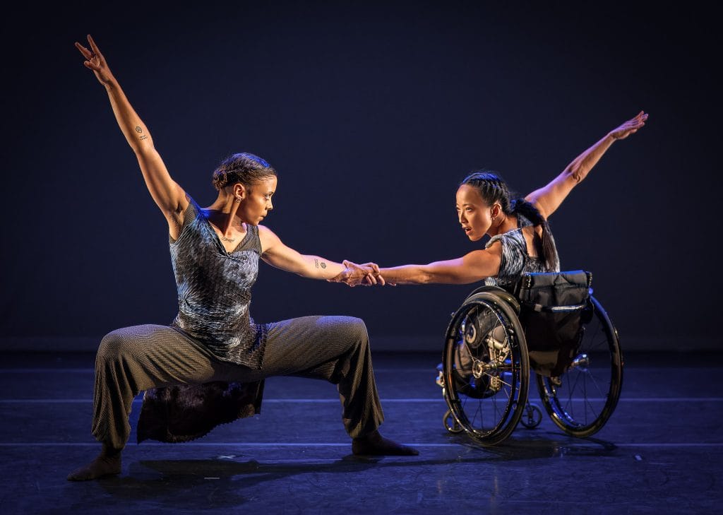 Dancers Alaja and Julie grasp each other's forearms in counterbalance with fierce expressions in their faces. Alaja holds a deep plie in second position while Julie reaches with one arm out from her wheelchair. 