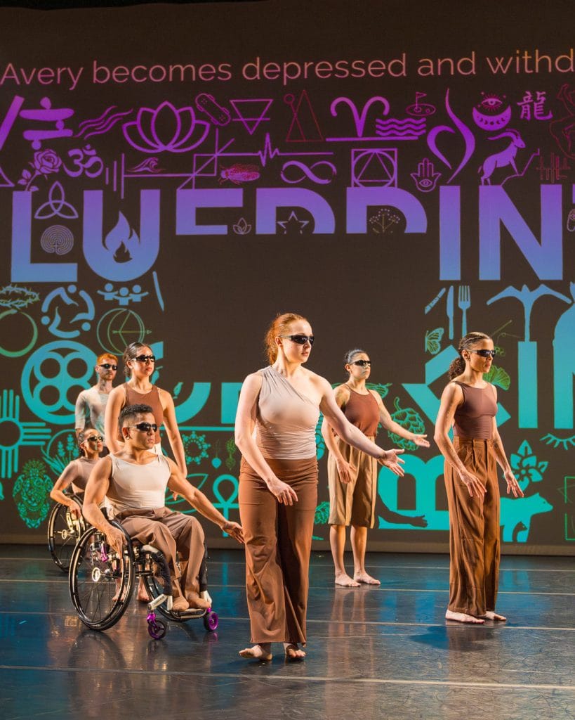 Seven disabled and non-disabled dancers gesture by raising one upper-right limb, wearing sunglasses and tan/brown costumes. Projected in the background is an abstract image composed of white symbols and text that reads "Blueprints of" with captions above this reading "Avery becomes depressed and withdrawn."