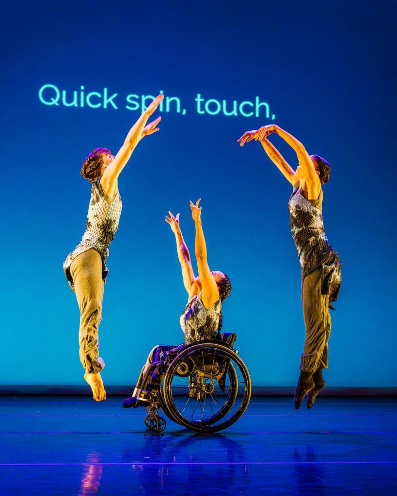 In the center, dancer Julie reaches two arms up from her wheelchair, while dancers Anna and Alaja leap up and away from Julie. On a blue lit background, text reads "quick spin, touch."