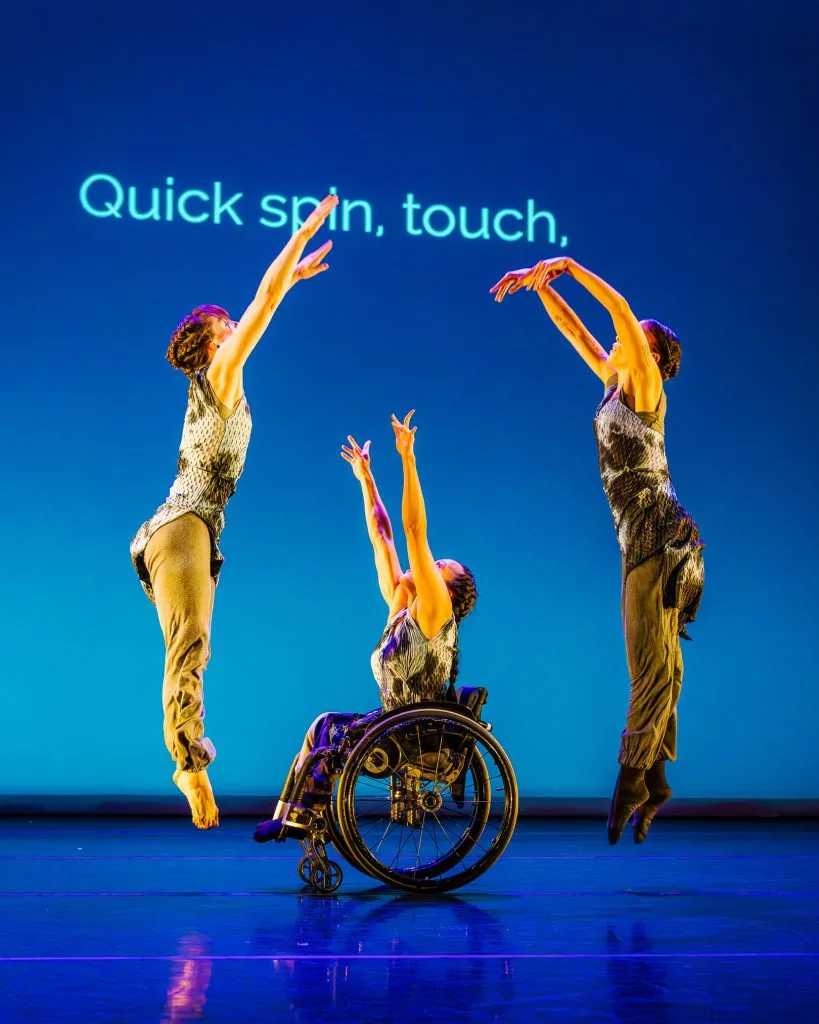 In the center, dancer Julie reaches two arms up from her wheelchair, while dancers Anna and Alaja leap up and away from Julie. On a blue lit background, text reads "quick spin, touch."