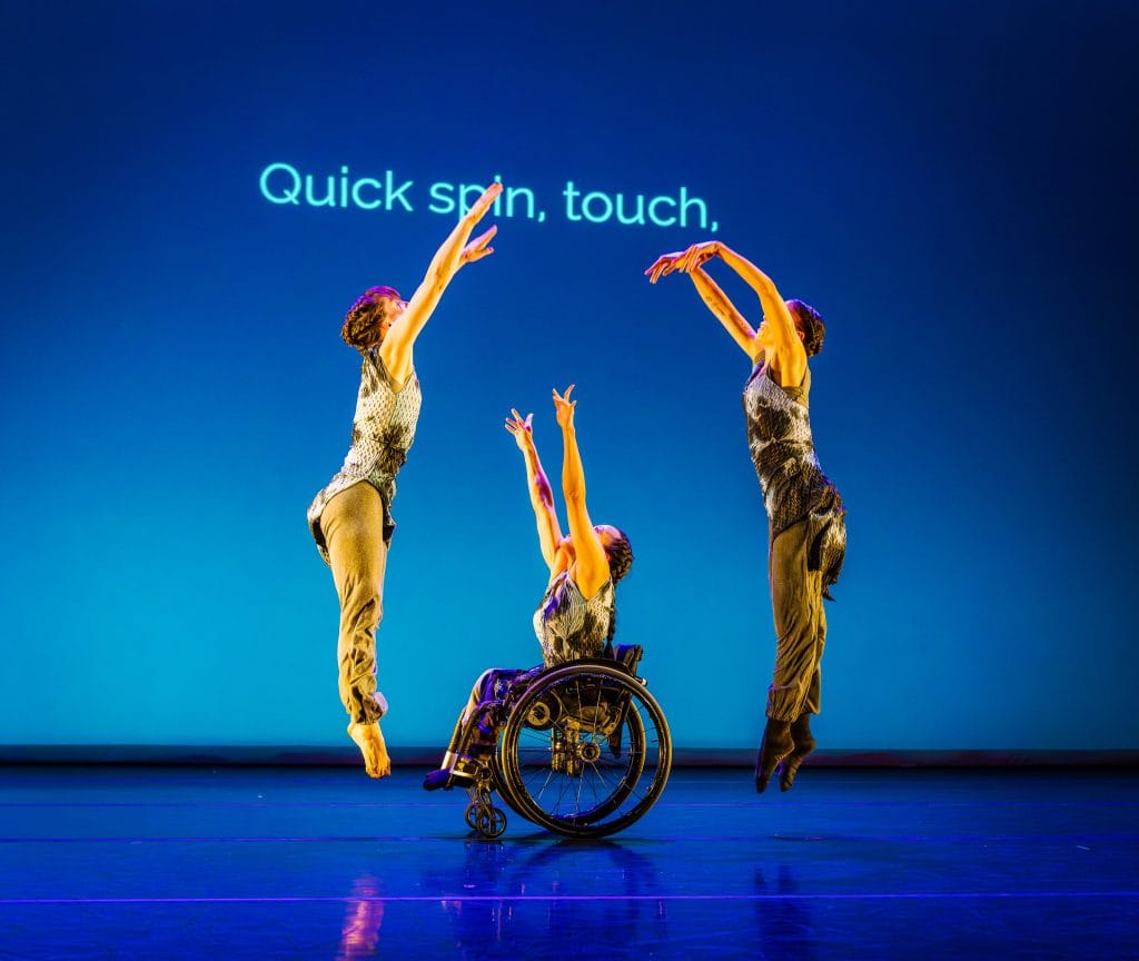 In the center, dancer Julie reaches two arms up from her wheelchair, while dancers Anna and Alaja leap up and away from Julie. On a blue lit background, text reads "quick spin, touch."