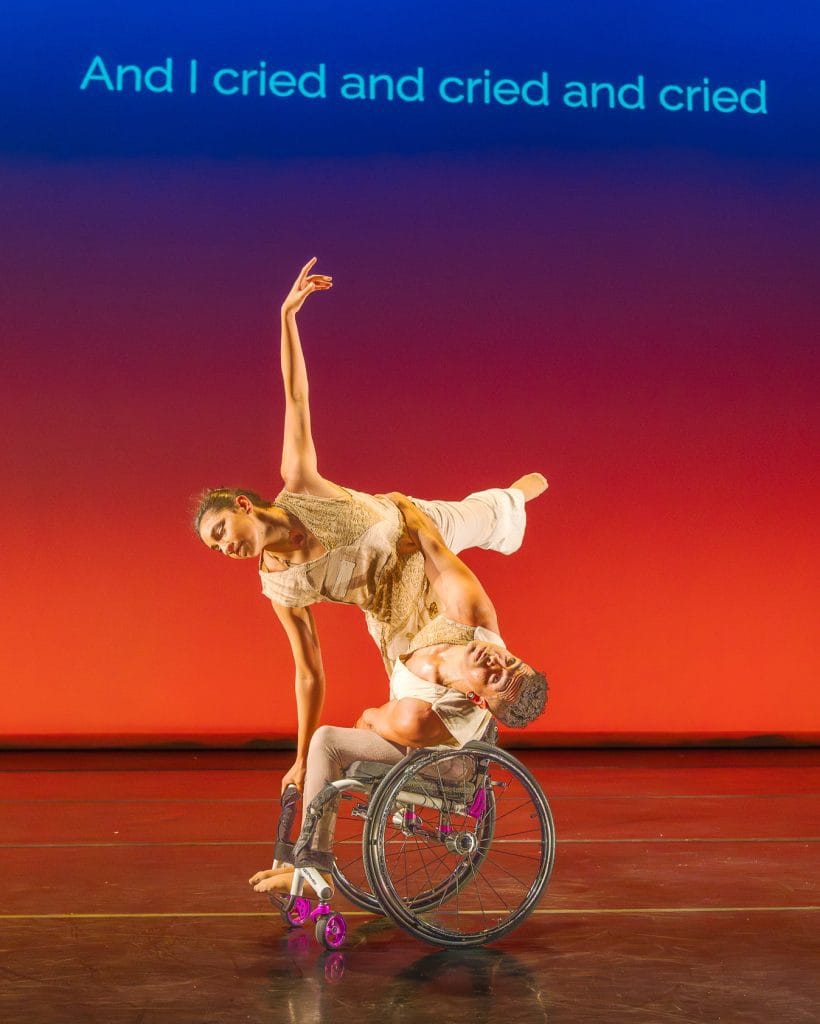 JanpiStar leans their torso to one side in their wheelchair, as Zara does an arabesque with one hand on the chair for support and stability. Against an orange - blue gradient, subtitles read "And I cried and cried and cried."