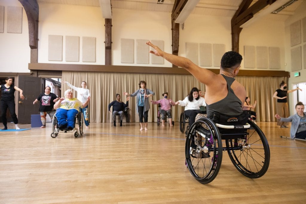 JanpiStar, who is a wheelchair user, teaches a dance class. They extend one limb in an upwards diagonal with sitting and standing dancers follow.