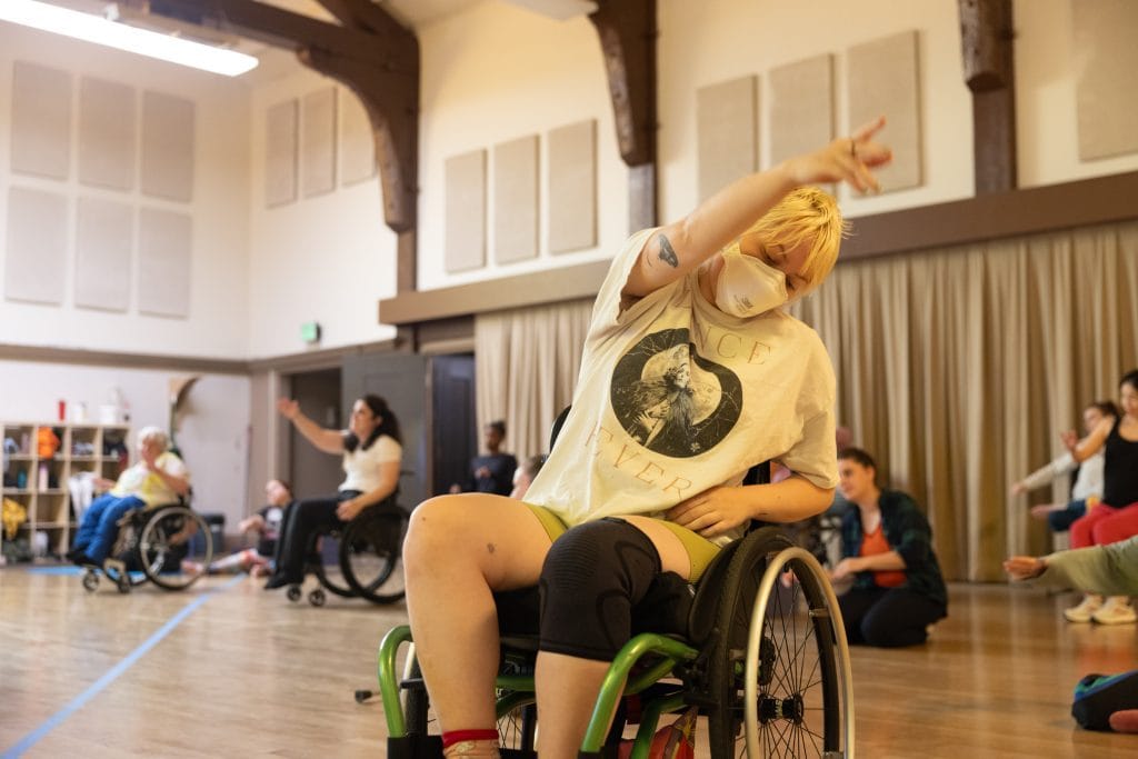 A Summer Intensive participant, who uses a wheelchair, reaches to the side in a torso lunge with an upper limb extended.