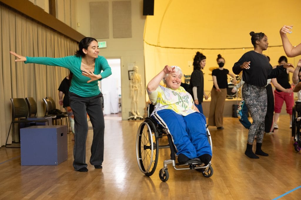 During a salsa class, two summer intensive participants smile and extend their limbs while others improvise in the background. One dancer uses a wheelchair, the other is standing.