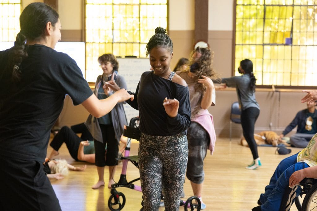 A diverse group of disabled and non-disabled dancers improvise in a dance studio, smiling.