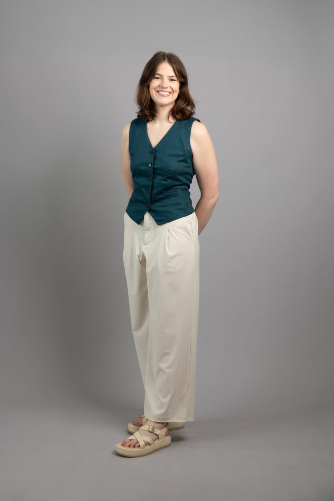 A portrait of Alannah, a tall white woman with mid-length wavy brown hair, wearing a jade vest. She stands with her arms behind her back.