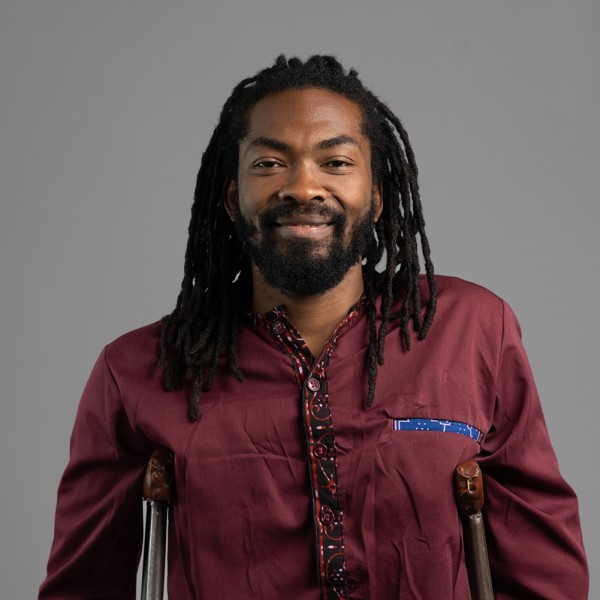 2025AXIS_RobSuguitan_TebandekeHeadshot Tebandeke poses for the camera with a smile. He is dark brown skinned African with black dreadlocks on his head, wearing a red collared shirt and standing with crutches supporting his upper body.