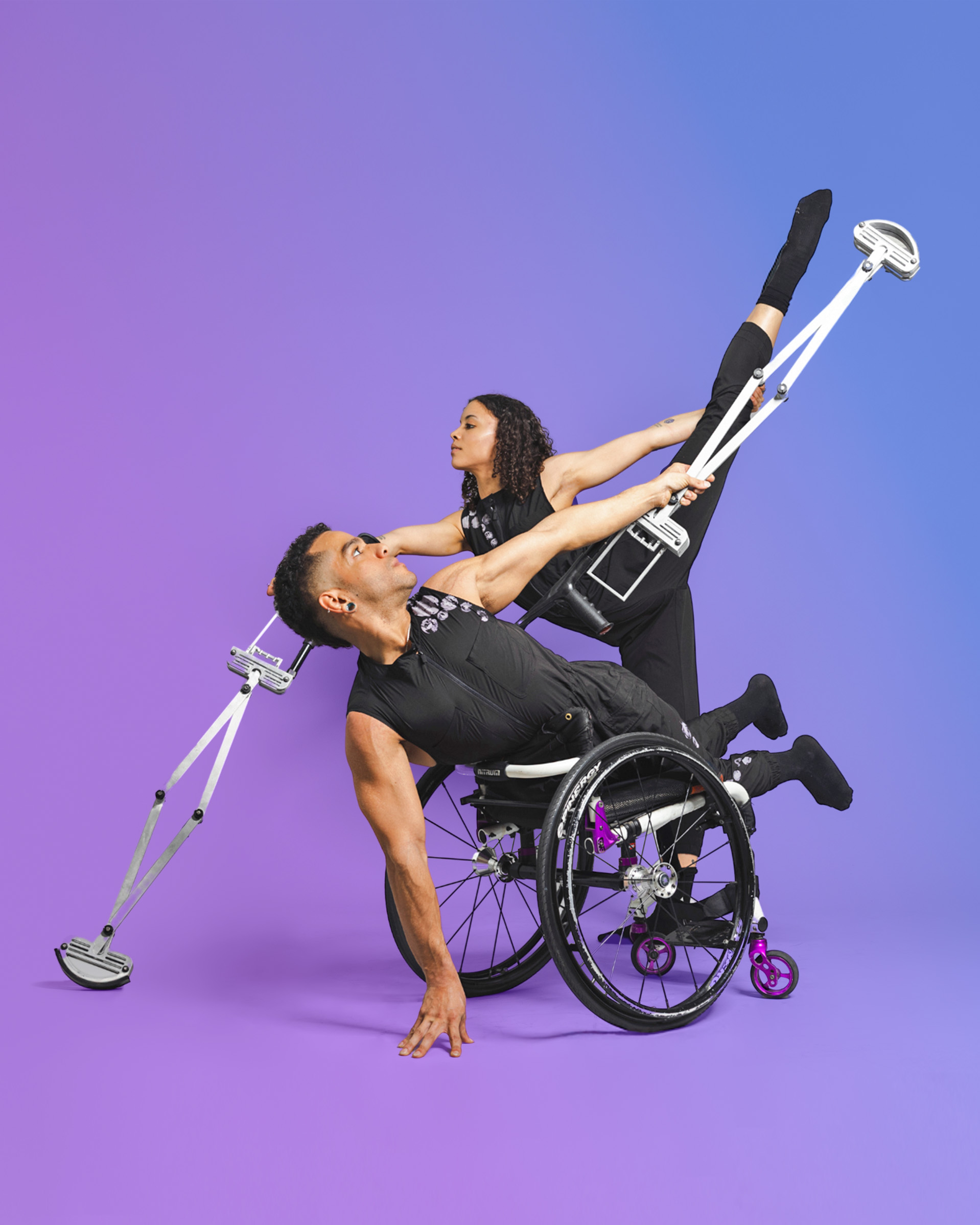 In the foreground, AXIS Dancer JanpiStar lies backwards and belly-forwards on top of their manual wheelchair, holding one arm on the ground as they twist their torso upwards to extend one long, silver telescoping crutch into the air. Behind them, Alaja mirrors this gesture by placing one telescopic crutch on the ground; using this to support their weight as they lift one leg up into a high arabesque that is parallel to Janpi’s crutch.