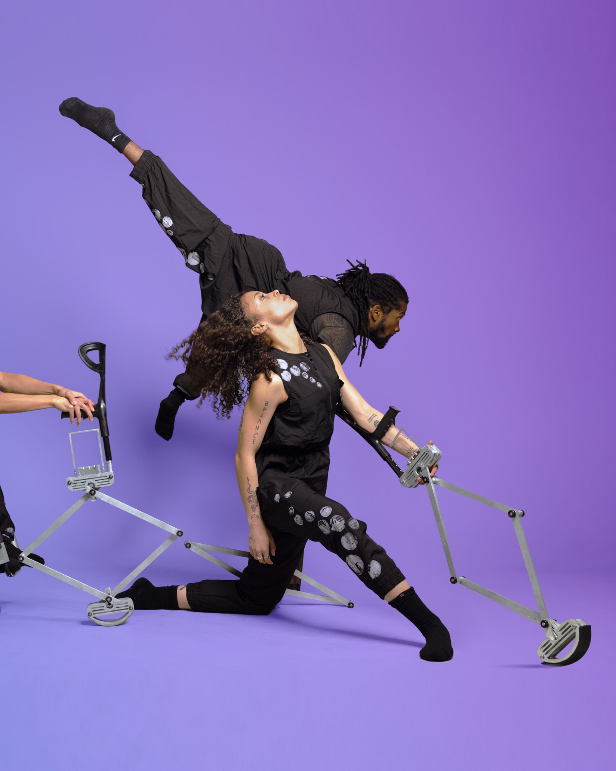 AXIS Dancers Julie and Alaja lean forward as they each hold out one silver, triangular telescoping crutch. Julie sits in her manual wheelchair; Alaja places one knee down in a lunge. Behind them, Tebandeke balances on his two crutches as he lifts one leg into a high arabesque; his other leg points towards the ground.