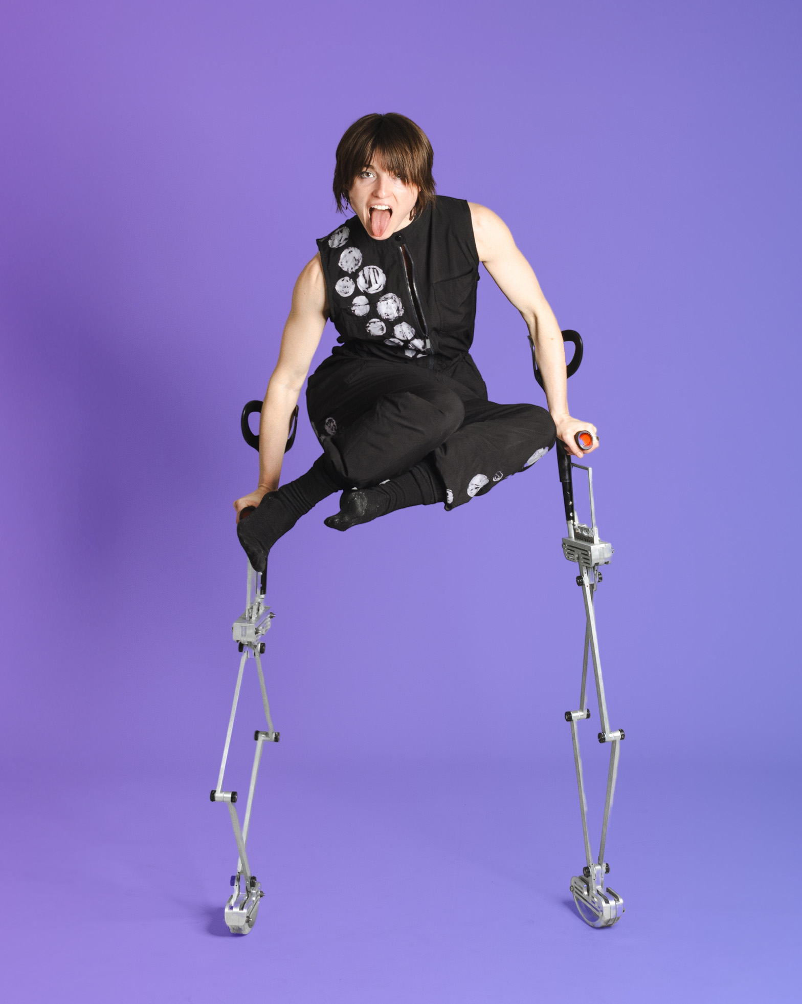 AXIS Dancer Anna balances on two silver telescopic forearm crutches as she lifts her body high in the air, bringing her knees close to her chest. She wears a black jumpsuit, and sticks her tongue out with a fierce and whimsical expression.