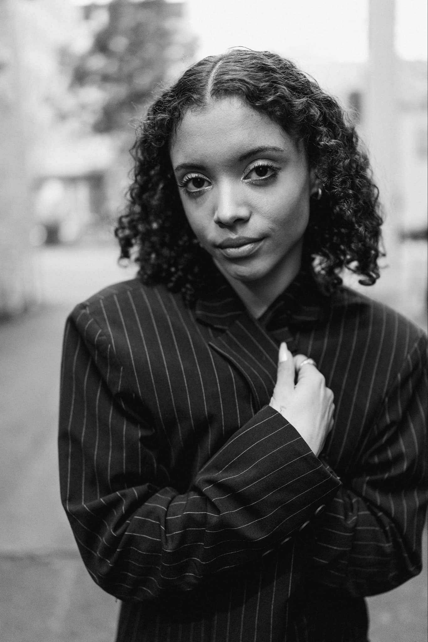 A(laj)a Badalich is black/mixed presenting genderfluid human, who uses they/she pronouns. They have brown curly hair, brown skin, and brown big eyes. In this black and white image they look directly at the camera and with their arms crossed, they use the lapels to pull a pinstripe suit jacket closed across their chest.