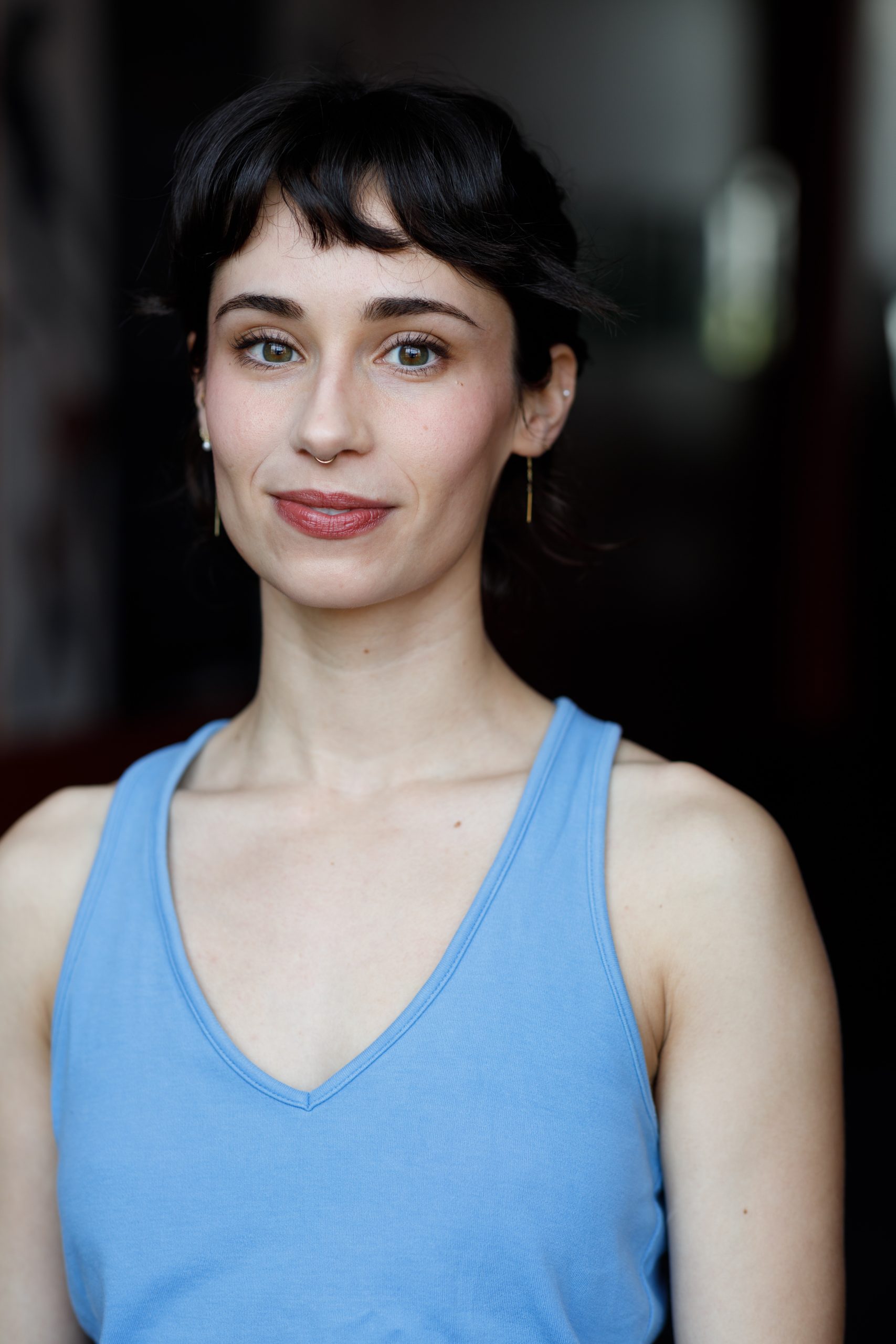 Clairey's headshot is photographed against a dark, blurred background. She has dark, Mediterranean features, hazel eyes, and pale skin. She is wearing a light blue tank top, gold jewelry in her ears and septum. She has a slight smile.