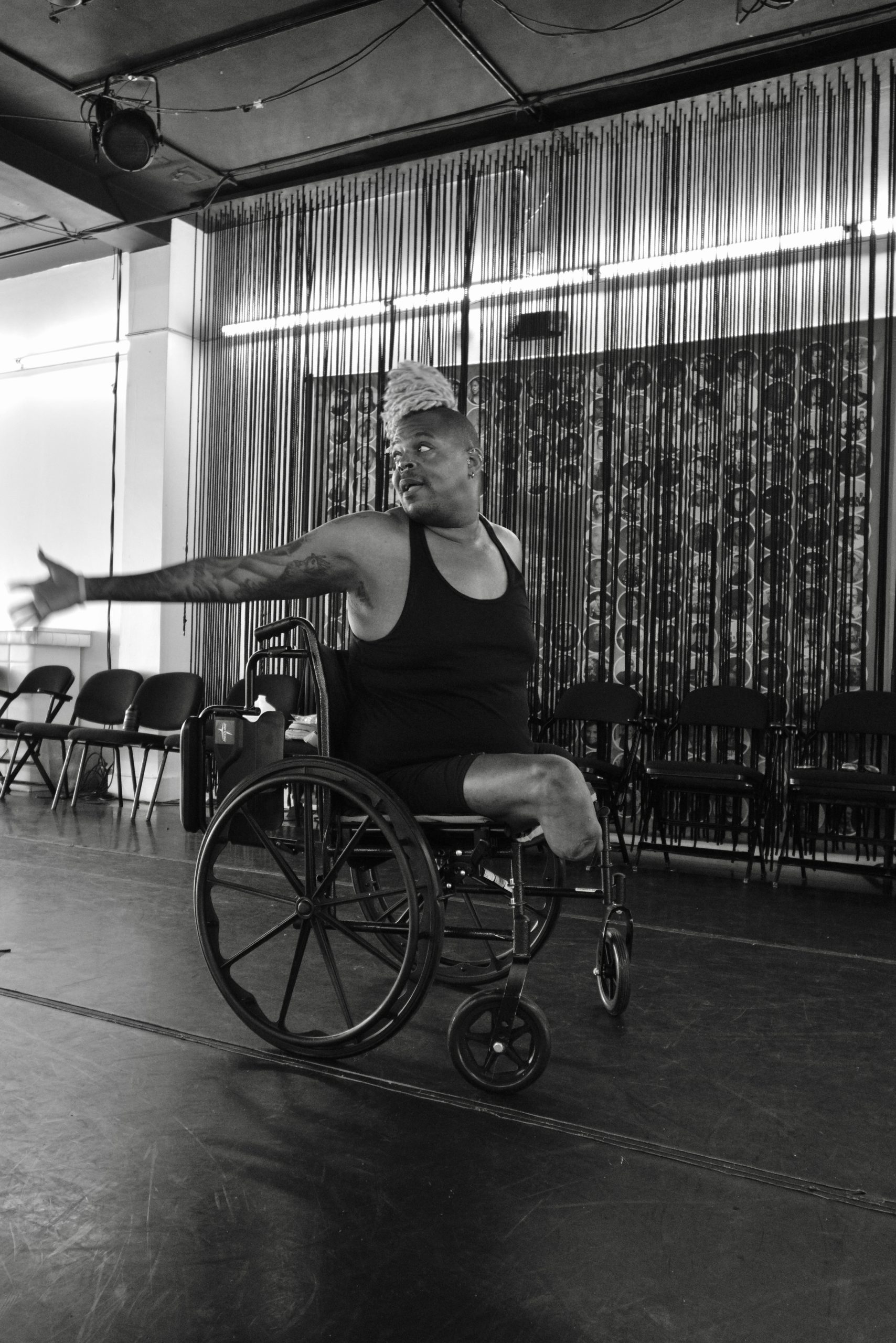 A black-and-white image of Hector, an Afro-Latino brown skin non-binary transfemme double amputee who’s right leg is below the knee & has a left above the knee in a wheelchair, engaged in a dance rehearsal. They wear a sleeveless top and have light-colored dreadlocks tied up. They are in a rehearsal space with chairs lined up in the background and vertical strings or wires forming part of the décor. Their posture and outstretched arms convey a sense of movement and emotion.