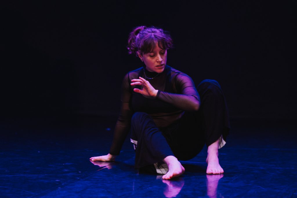 A dramatically lit photo depicts Hannah in an almost seated position preparing to propel herself forward with the leading elbow bent and focused towards the floor, which glows a subtle blue. She's wearing flowing black pants with a white stripe down the side, and a long sleeved black mesh top. Her expression is focused and soft.