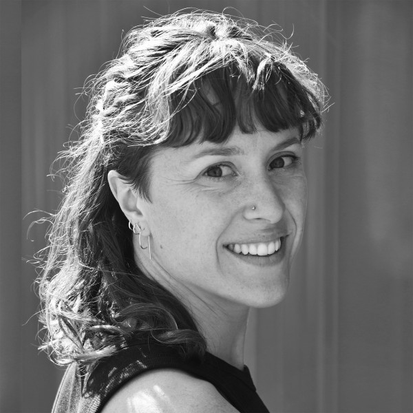 HannahWestbrookPhoto A black and white photo depicts Hannah looking over her right shoulder and smiling towards the viewer. She's a white woman with wavy light brown hair cut into bangs and falling around shoulder length. She is wearing a tank top and has small silver jewelry accenting her ears and nose.